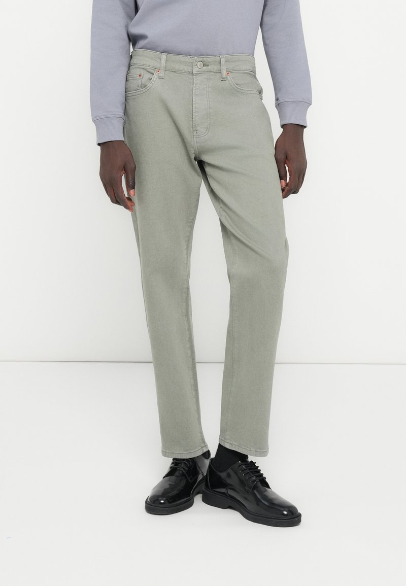 Φωτεινά πράσινο βαμβακερό παντελόνι με ίσια γραμμή, τσέπες, πάνω κουμπί και θηλιές για ζώνη, συνδυασμένο με μαύρα παπούτσια και γκρι φούτερ.