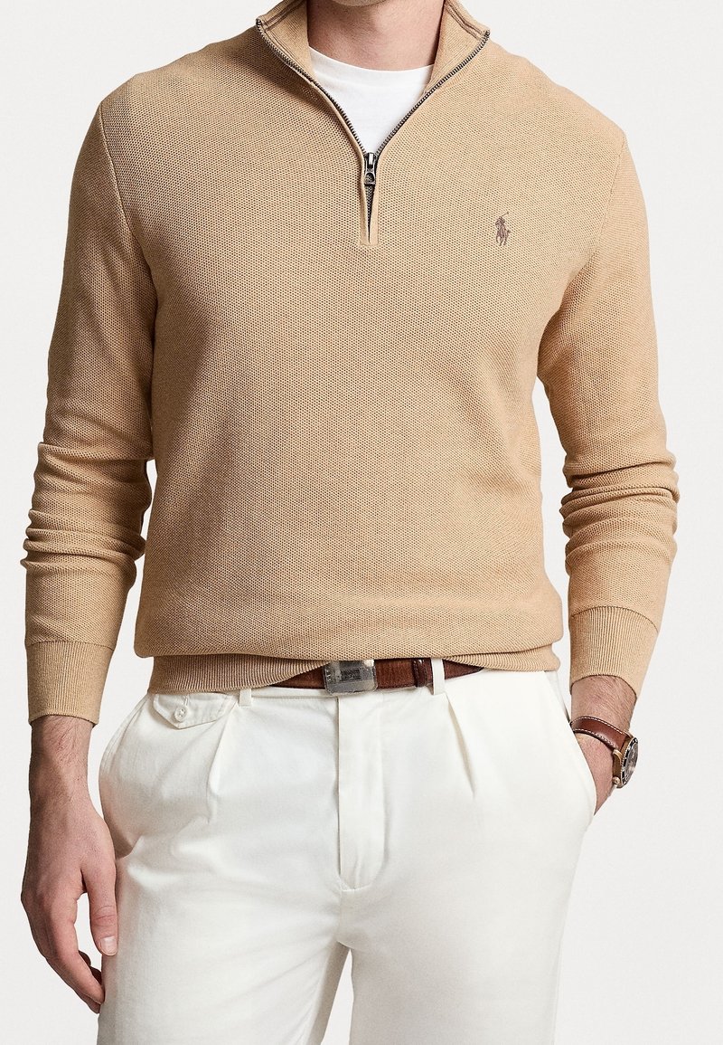 Homme portant un pull en maille beige avec demi-fermeture éclair, un t-shirt blanc, un pantalon blanc plissé, une ceinture en cuir marron et une montre-bracelet avec un bracelet en cuir marron.