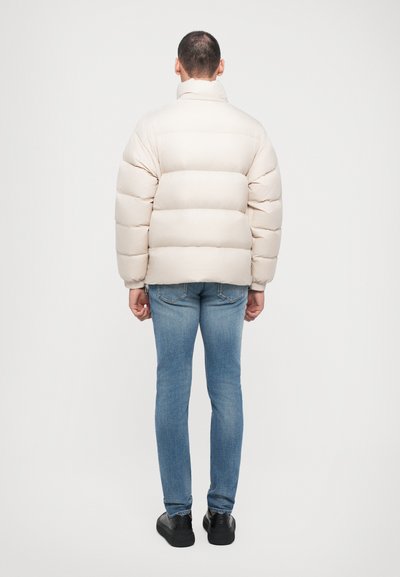 Beige pufferjakke med høj krave, udstyret med en quilted design, tykk isolering og ribbede manchetter, parret med blå jeans og sorte sko.