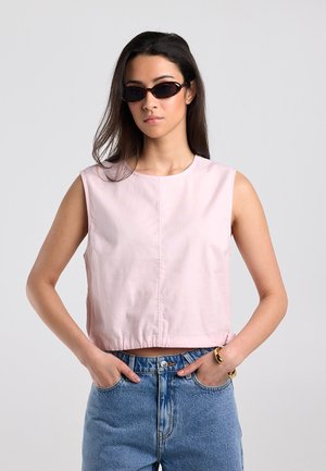 Vrouw met zwarte ovale zonnebril, mouwloze lichtroze top, blauwe spijkerbroek en gouden armbanden, staand met handen in de zakken.