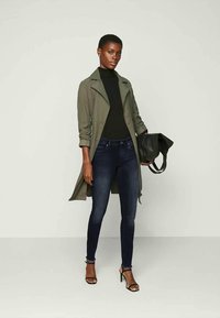 Trench coat caqui com um corte ajustado, suéter preto, jeans skinny azul-escuro, mala preta e sapatos de salto com tiras em uma modelo contra um fundo neutro.