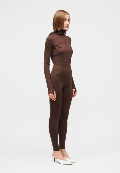 Rudos spalvos ilgarankovė bodžio tipo bluzė su gaubtu, aptempto silueto, su užtrauktukais kišenėse ir lygiu paviršiumi, derinama su baltais mulei tipo batais.