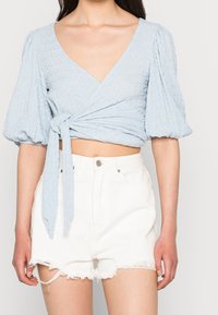 Top avvolgente blu chiaro, stropicciato, con maniche a sbuffo, legato in vita. Abbinato a pantaloni corti bianchi strappati, con orli sfrangiati e tasche.
