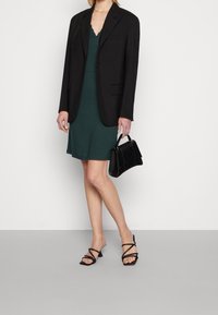 Kobieta w ciemnozielonej sukience z falbaniastym wykończeniem, czarnym żakiecie, czarnych sandałach na obcasie, trzymająca małą czarną torbę na tle białym.