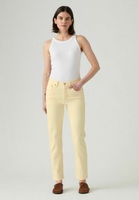 Vaqueros amarillos claros con un diseño ajustado, estilo de cinco bolsillos y costuras en contraste. Combinados con una camiseta sin mangas blanca y mocasines marrones.