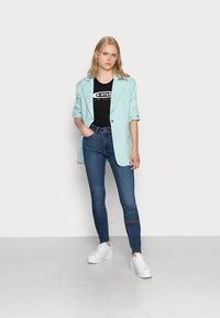 Blazer verde menta sobre uma t-shirt preta, combinado com jeans skinny azul escuro e sapatilhas brancas de plataforma, colocado contra um fundo neutro.