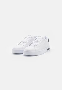 White sneakers made of textured synthetic material with a smooth toe design, flat laces, and a black accent at the heel.