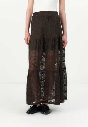 Kvinde iført en mørkebrun, gennemsigtig maxi nederdel i blonder med lagdelt design, kombineret med sorte snørebåndssko og en hvid top.