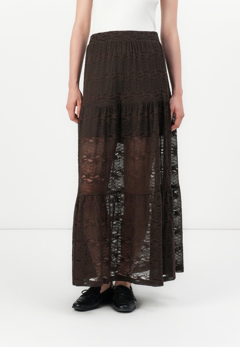 Femme portant une jupe longue en dentelle marron foncé transparente avec un design à étages, associée à des chaussures noires à lacets et un haut blanc.