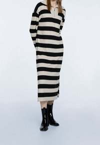 Abito a righe in nero e beige, con colletto mezza zip, maniche lunghe, silhouette oversize e spacchi laterali. Indossato con stivali neri lucidi.
