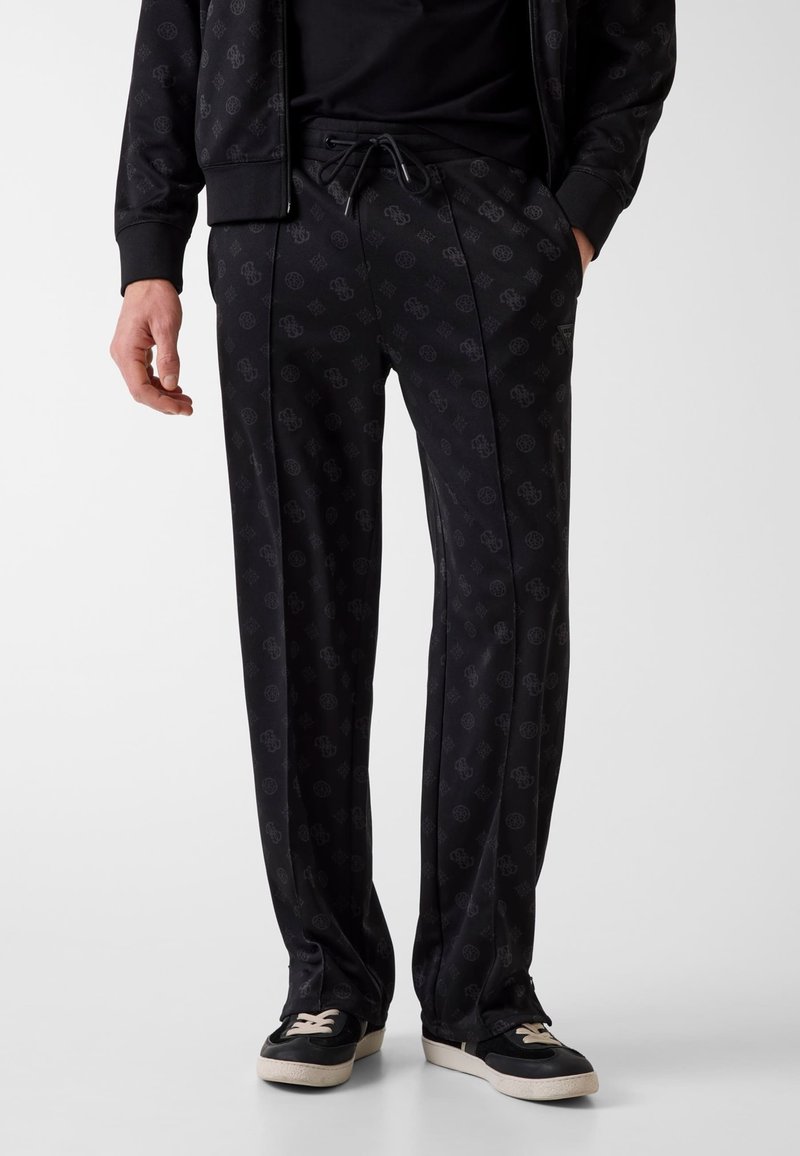 Uomo che indossa pantaloni con coulisse neri a motivo, giacca coordinata e scarpe da ginnastica nere con suole e lacci bianchi, in piedi davanti a uno sfondo semplice.