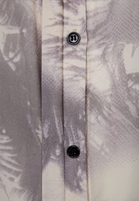 Leichte Bluse aus Stoff mit einem subtilen grauen und cremefarbenen Palmenmuster. Verfügt über eine durchgehende Knopfleiste mit runden schwarzen Knöpfen und einer glatten Textur.