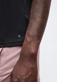 Gros plan sur un avant-bras à la peau foncée tatoué, portant une chemise noire texturée et un pantalon rose clair sur un fond blanc.
