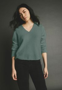 Maglione verde acqua a V, realizzato in un materiale morbido, con maniche lunghe e orlo corto, abbinato a pantaloni neri a trama.
