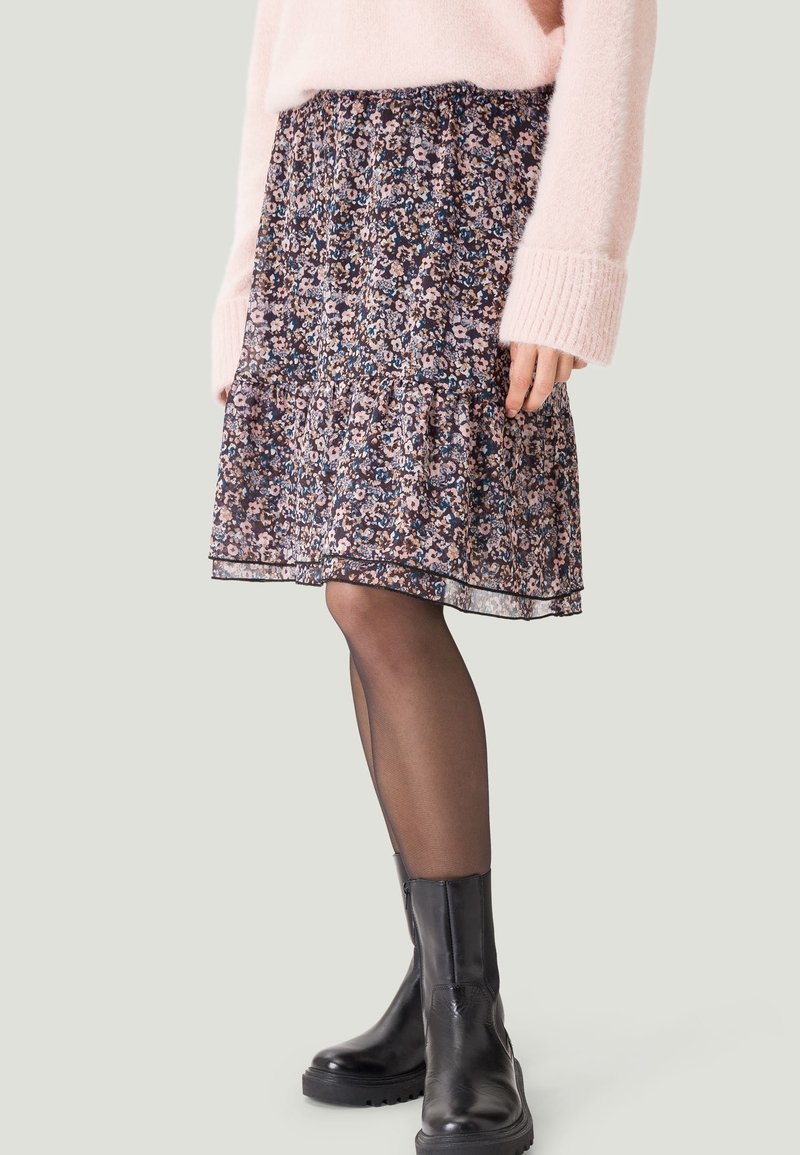 Femme portant une jupe à motifs floraux longueur genou, des collants noirs transparents, des bottines en cuir noires et un pull en tricot rose clair.