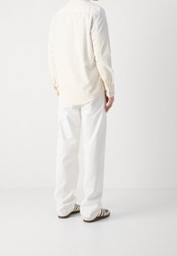 Camicia button-up color crema con una texture morbida, abbinata a pantaloni bianchi a vestibilità ampia e scarpe casual a righe marroni. Dettagli di design sottili visibili.
