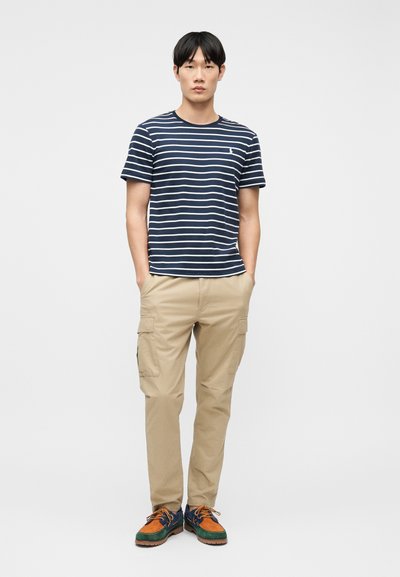 Νέος άνδρας που στέκεται με τα χέρια στις τσέπες, φορώντας μπλε ναυτικό και λευκή ριγέ μπλούζα, μπεζ παντελόνι cargo και πολύχρωμα παπούτσια με κορδόνια σε λευκό φόντο.