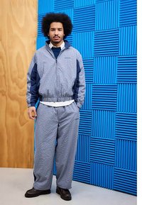 Windbreaker and matching pants in blue and gray houndstooth pattern, with a navy accent. Smooth fabric; relaxed fit. Dark shoes.