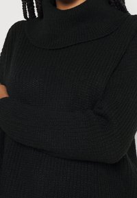 Pull noir en tricot avec un col roulé haut et un tissu texturé. Manches longues avec une coupe ample, présentant des détails côtelés au niveau du col.