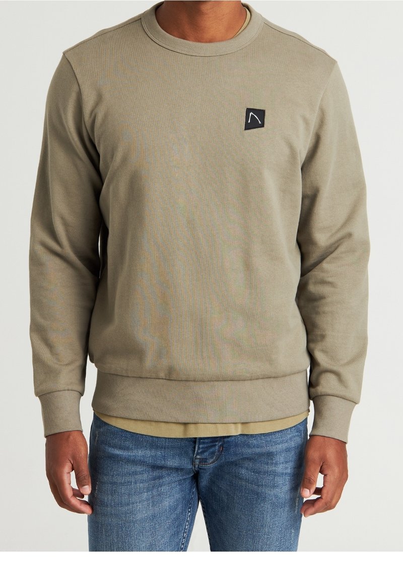 Felpa khaki, colletto a giro, polsini e orlo a costine, piccolo logo nero ricamato sul petto, texture morbida, abbinata a jeans blu.
