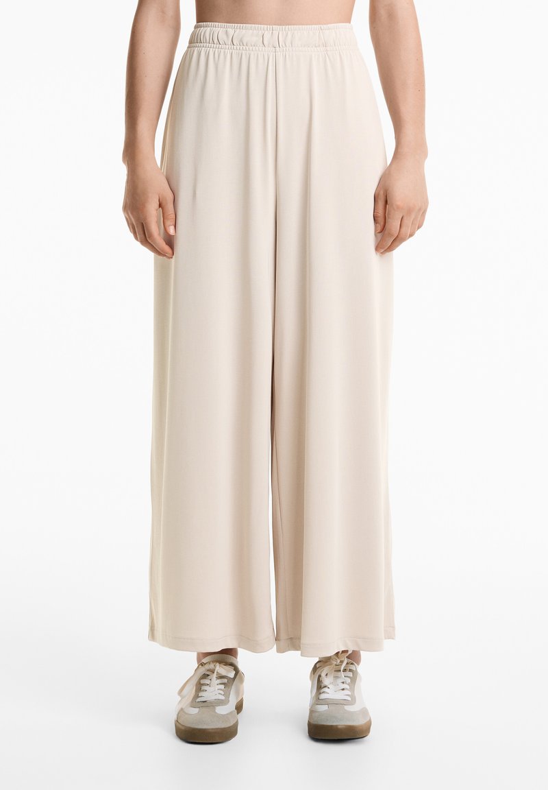 Wide-leg beige pants made of soft fabric, featuring an elastic waistband. Worn with light gray and beige sneakers.