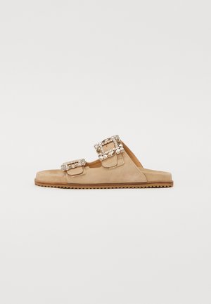 Sandalia deslizante de ante en beige con dos hebillas decorativas que presentan acentos transparentes. Suela plana con un patrón de agarre texturizado. Diseño casual.