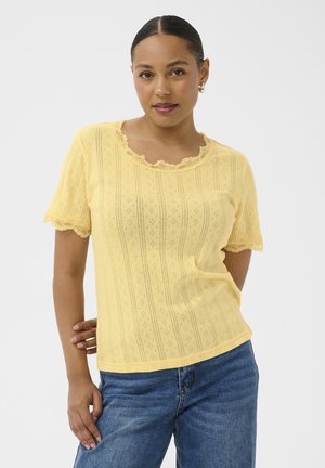 Femme aux cheveux foncés portant un haut à manches courtes orné de dentelle jaune et un jean bleu, debout avec une main derrière le dos.