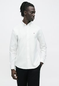 Homme aux dreadlocks portant une chemise blanche boutonnée avec un petit logo sur la poitrine et un pantalon noir, debout avec une main dans la poche.