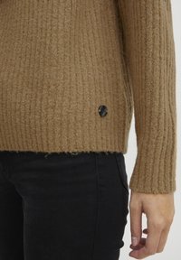 Pulover ribbed de culoare maron deschis, realizat dintr-un material texturat, având o fanta pe lateral și un accent cu un nasture negru aproape de tiv. Asortat cu blugi negri.