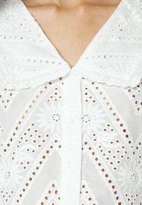 Blouse blanche avec un large col festonné et une broderie ajourée florale et géométrique complexe sur un tissu semi-transparent.