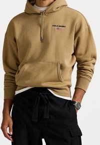 Mand iført tan Polo Sport-hoodie med amerikansk flaglogo, sorte cargobukser og et mørkt ur på venstre håndled.