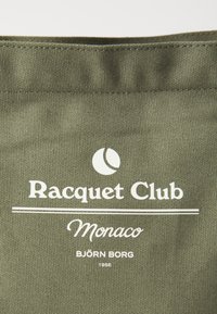 Olivovo zelená taška s bielym textom: "Racquet Club Monaco BJOR̈N BORG 1956." Text je potlačený v strede s kruhovým logom nad ním.