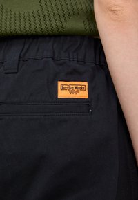 Black trousers with a textured fabric, featuring a visible orange label that reads "Service Works." Includes a front pocket with stitching detail.