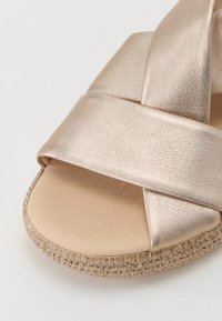 Metallic gold strappy sandal with crossed upper design, textured leather finish, and a flat woven sole. Smooth inner lining.