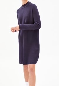 Personne portant une robe tricotée violet foncé à manches longues, longueur genou, avec un col montant, se tenant devant un fond blanc uni.