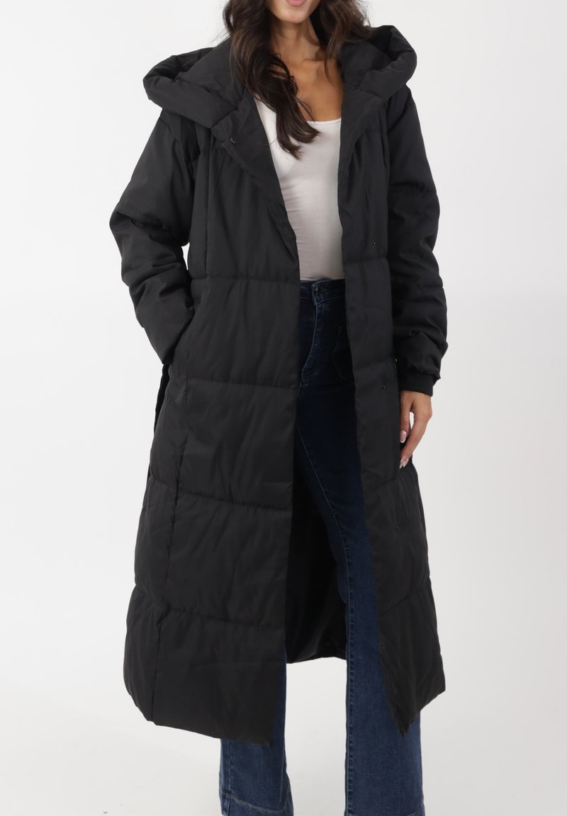 Piumino nero lungo con cappuccio, caratterizzato da un design trapuntato, tasche frontali e una vestibilità comoda, indossato sopra una maglietta bianca e jeans scuri.