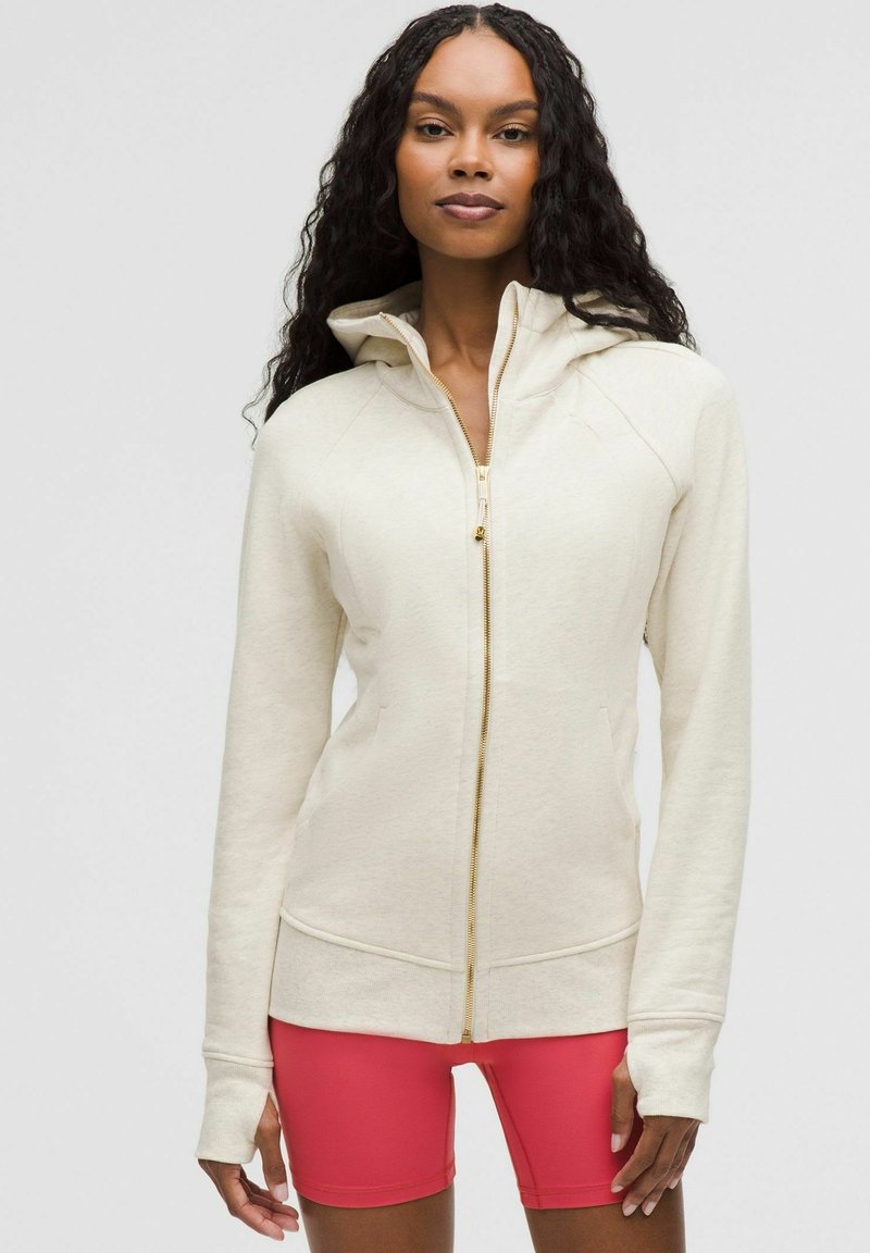 Svijetlo bež dukserica s patentnim zatvaračem izrađena od mekog materijala, s kapuljačom, raglan rukavima i zlatnim patentnim zatvaračem, u kombinaciji s ružičastim sportskim kratkim hlačama.