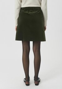 Donkergroene A-lijn rok met een gladde textuur, elastische tailleband en een knielange snit, gedragen met zwarte panty’s en schoenen.