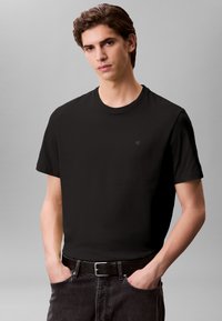 T-shirt preta de algodão com decote redondo, mangas curtas e um pequeno logótipo no canto superior esquerdo. Combinada com jeans de ganga escura e um cinto preto.