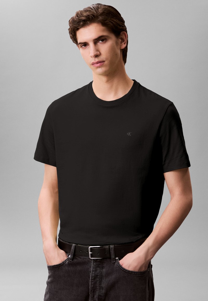 T-shirt preta de algodão com decote redondo, mangas curtas e um pequeno logótipo no canto superior esquerdo. Combinada com jeans de ganga escura e um cinto preto.