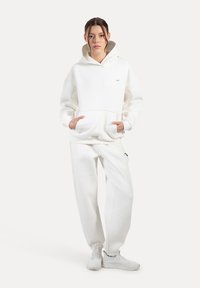 Weißer Kapuzenpullover mit Kängurutasche, kombiniert mit weißen Jogginghosen. Weiches Material, lockere Passform, minimalistisches Design, dezentes Branding-Detail.