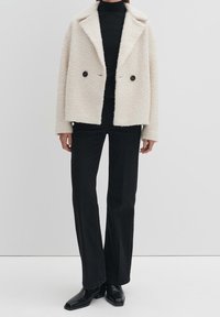 Cream-colored, textured, faux fur jacket with a collared neckline, double black button closure, and long sleeves paired with black wide-leg pants.