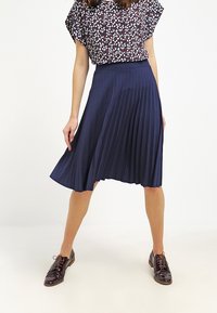 Marinblå plisserad kjol med en jämn textur; hög midja, knälång, matchad med en blus med blommönster och mörka oxfordskor.