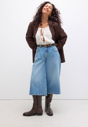 Mujer con cabello rizado sonríe, lleva una blazer marrón oscuro, blusa blanca con lazo en la parte delantera, vaqueros azules de pierna ancha, cinturón negro y botas marrón oscuro hasta la rodilla.