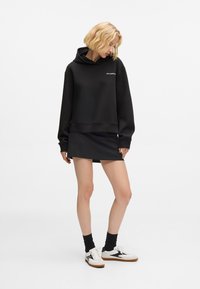 Mulher loira vestindo um hoodie preto, saia mini preta, meias pretas e tênis brancos, de pé contra um fundo branco simples.