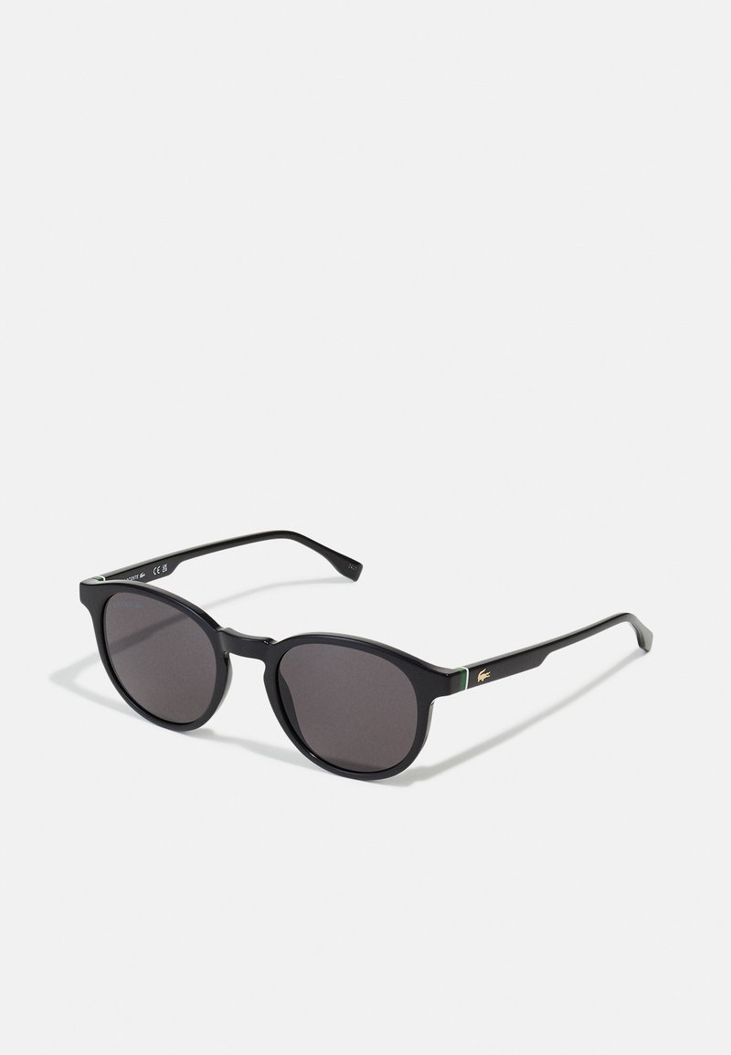 Ochelari de soare negri cu rame rotunjite, lentile gri închis și brațe negre elegante, având un mic accent logo verde și aurie.