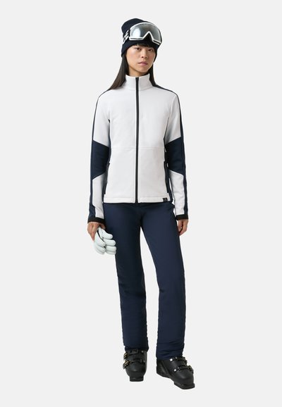 Weiße und marineblaue Skijacke mit Reißverschluss, strukturiertem Stoff, kombiniert mit marineblauer Skihose, schwarzen Skischuhen und einer schwarzen Mütze mit Skibrille.