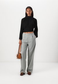 Top lungo a maniche lunghe plissettato nero abbinato a pantaloni plissettati grigio chiaro a vita alta. Presenta una borsa a tracolla marrone e mocassini marroni con dettagli.