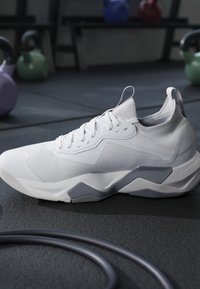 Scarpa sportiva bianca con dettagli grigi sulla suola, posta sul pavimento della palestra vicino a kettlebell e una fascia nera per esercizi arrotolata.