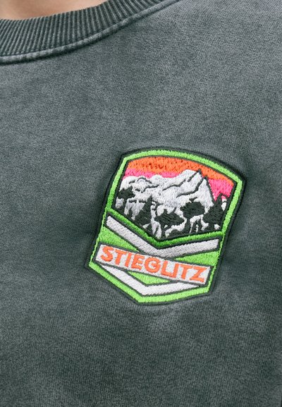 Tmavošedá mikina s farebnou vyšívanou nášivkou zobrazujúcou hory, stromy a slovo "STIEGLITZ" v oranžovej farbe, orámované zelenou.